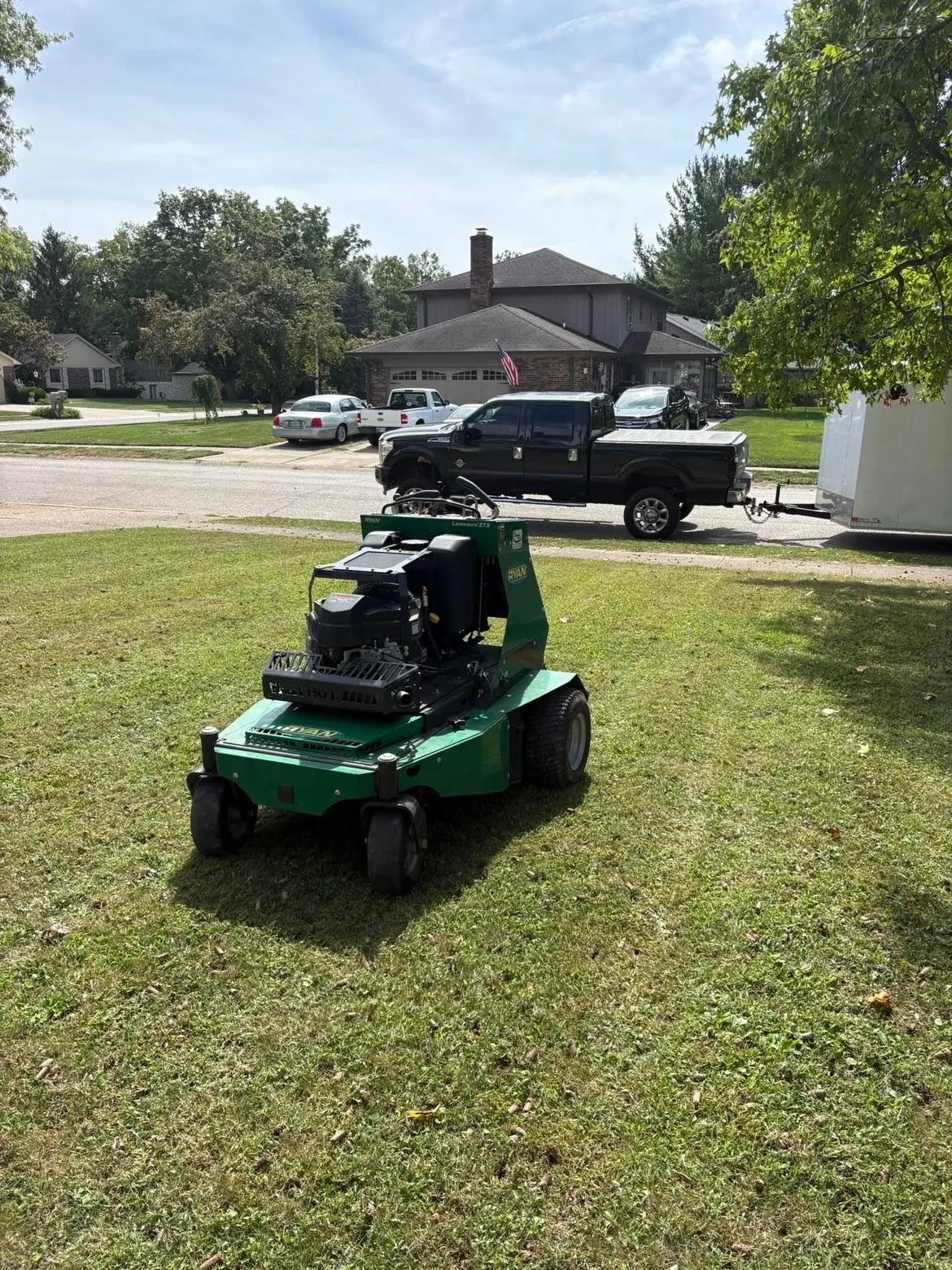 His aerator on a front lawn, the truck and trailer at the curb.